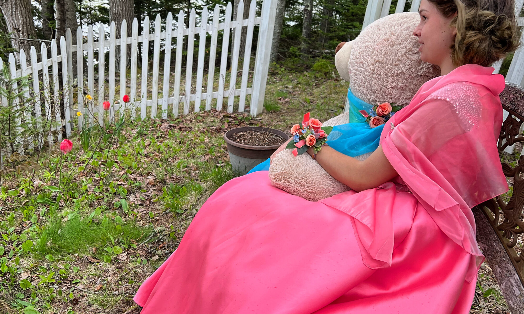 A Teenage Girl sits with life-size teddy bear in coral pink formal dress on a bench looking out to sea at Moo's Corner